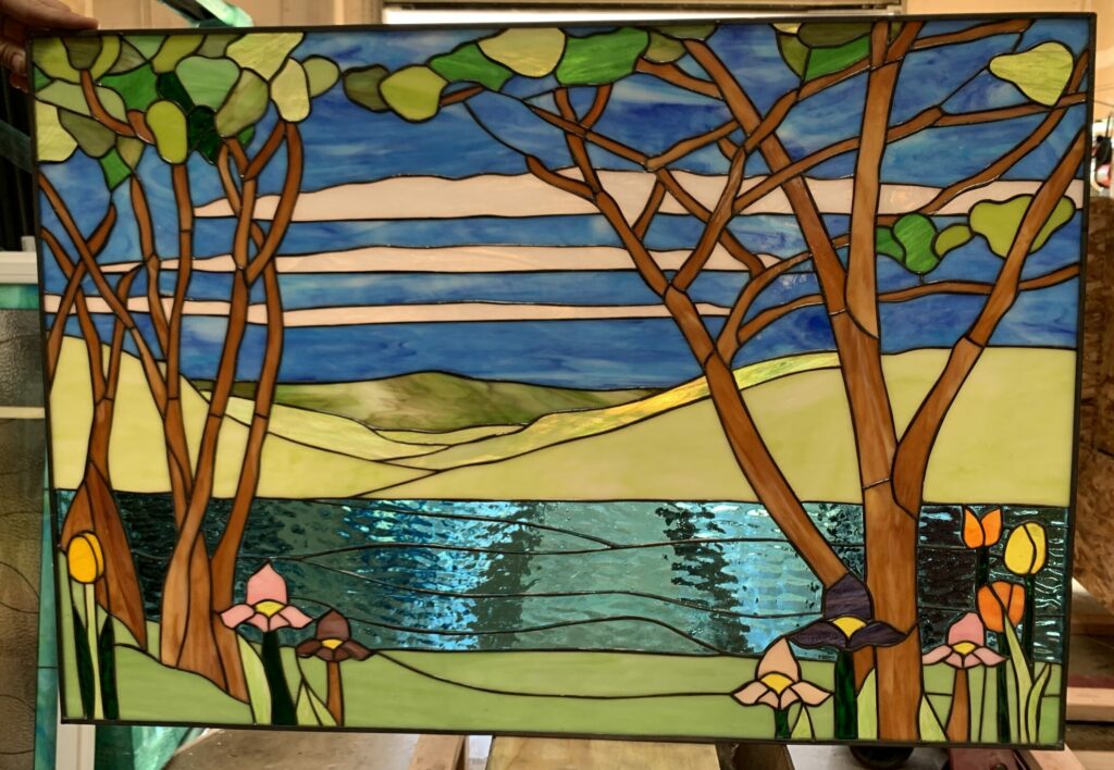 Rectangular stained glass panel depicting a vibrant landscape scene with two leafy trees framing a sparkling blue lake, rolling green hills in the background under a blue and white cloudy sky, and colorful foreground flowers.
