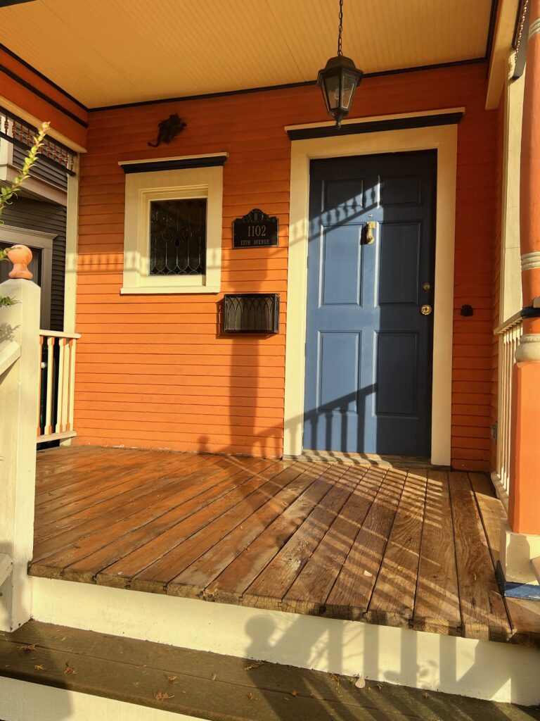 A small rectangular window with clear leaded glass featuring symmetrical scrollwork patterns, set in a vibrant orange house exterior next to a blue front door.