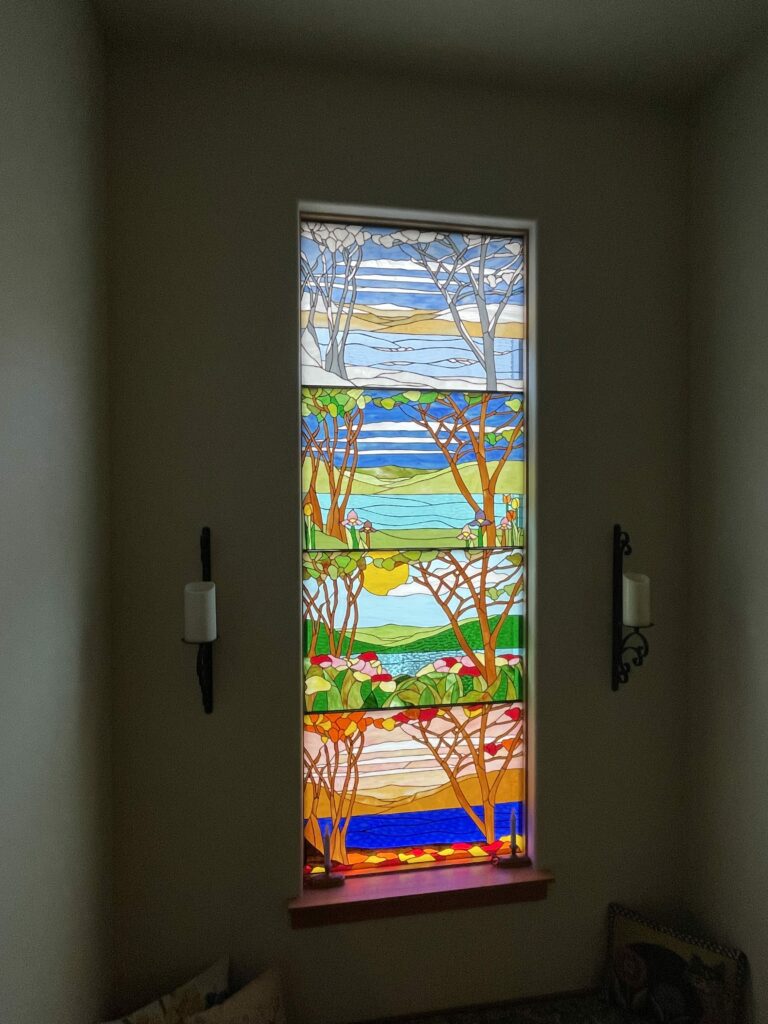 A tall, rectangular leaded stained glass window depicting the four seasons. From top to bottom: Winter with white and grey bare trees over a snowy landscape; Spring with green-leafed trees, irises, and tulips beside a blue lake; Summer with lush green trees, a bright yellow sun, and vibrant flowers; and Autumn with orange and red trees over a golden landscape and deep blue water.