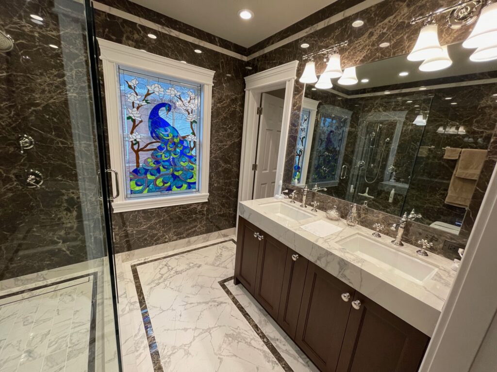 A vibrant stained glass window depicting a majestic blue and green peacock with a long, flowing tail, perched on a brown tree branch adorned with delicate white cherry blossoms. The leaded glass panel is installed in a white-framed window, enhancing a luxurious bathroom featuring dark brown marble walls, a white marble floor, and a double vanity with polished chrome fixtures.