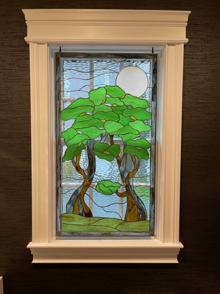 A rectangular stained glass window panel depicting a tranquil night scene with two stylized trees, featuring vibrant green leaves and dark brown, textured trunks. A large, clear white full moon is visible in the upper right. The background is made of wavy, light blue and clear textured glass, suggesting water, and a horizontal strip of textured green glass forms the ground. The panel is set within a white decorative frame against a dark, textured wall.