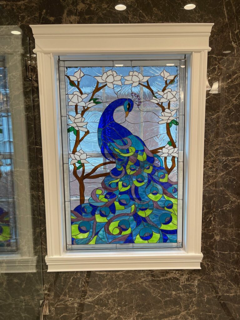 A rectangular, leaded stained glass panel in a white decorative frame, depicting a vibrant blue and green peacock perched on a brown branch with white magnolia flowers. The background glass is textured light blue, and the panel is set into a dark brown marbled wall.