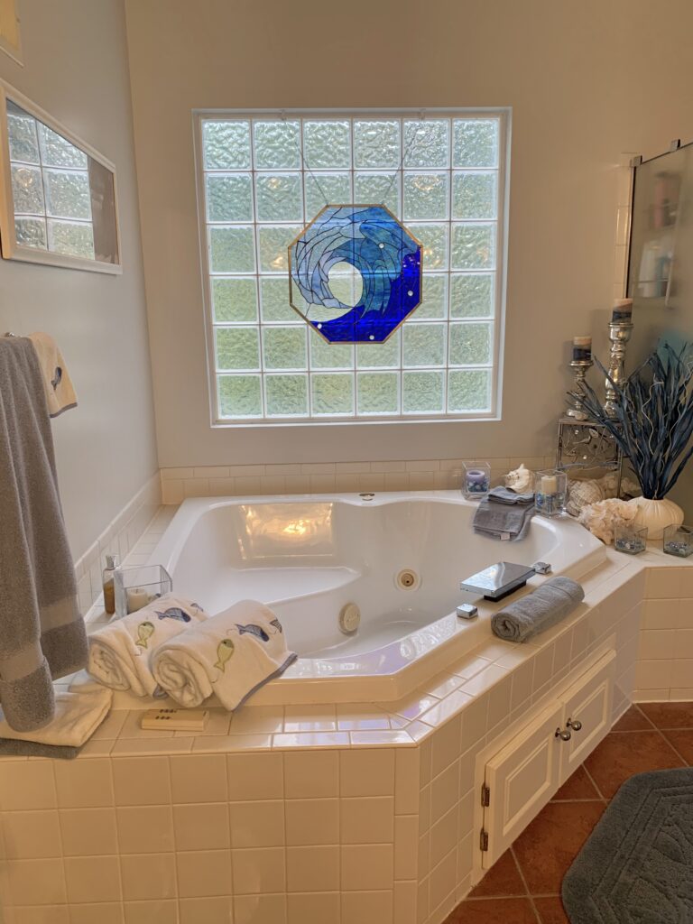 An octagonal stained glass window panel depicting a dynamic, swirling ocean wave in various shades of blue and white, with clear textured glass. The panel is framed in a brass-colored lead came and hangs in front of a frosted glass block window above a white jacuzzi tub in a bathroom setting.