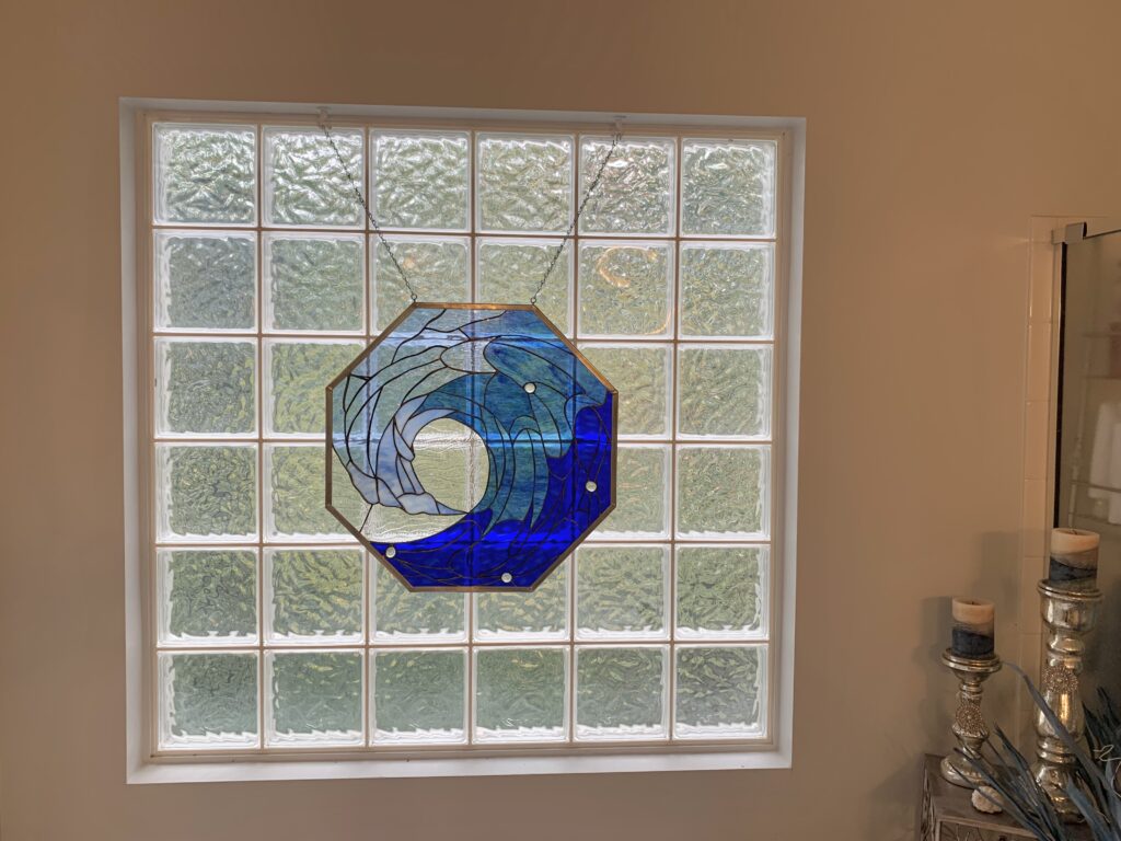 An octagonal stained glass panel featuring a dynamic ocean wave design in various shades of blue and white glass, framed in copper, hanging by chains against a textured glass block window.