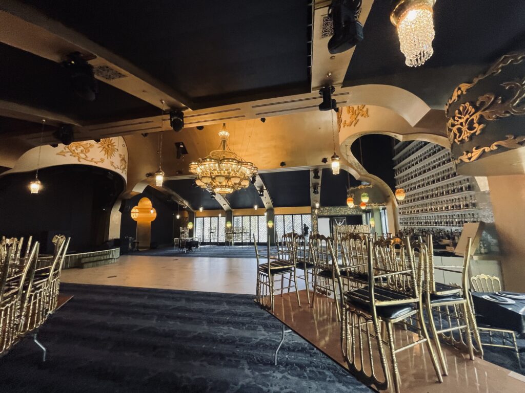A lavish event hall interior featuring a grand crystal chandelier, multiple colored art glass pendant lights, ornate gold and black ceiling details, stacked gold banquet chairs, and a dark patterned floor.