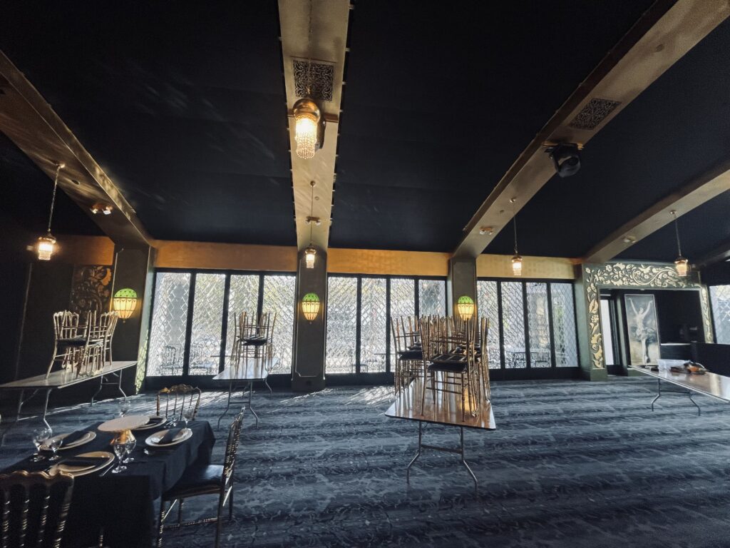 Interior view of an opulent event hall showcasing large windows with clear, diamond-patterned leaded glass panels. The room features a dark ceiling, golden ceiling beams, wall columns with decorative green glass sconces, and set tables with black linens and ornate gold chairs.