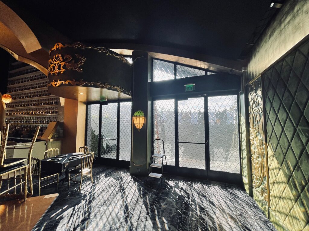 A luxurious interior scene featuring expansive window walls and doors crafted with diamond-patterned leaded glass, allowing bright sunlight to cast striking geometric patterns across the dark, reflective floor. The room is adorned with ornate gold decorative elements, dark green tufted velvet wall panels, and elegant dining furniture, highlighting a rich blend of light, shadow, and sophisticated design.