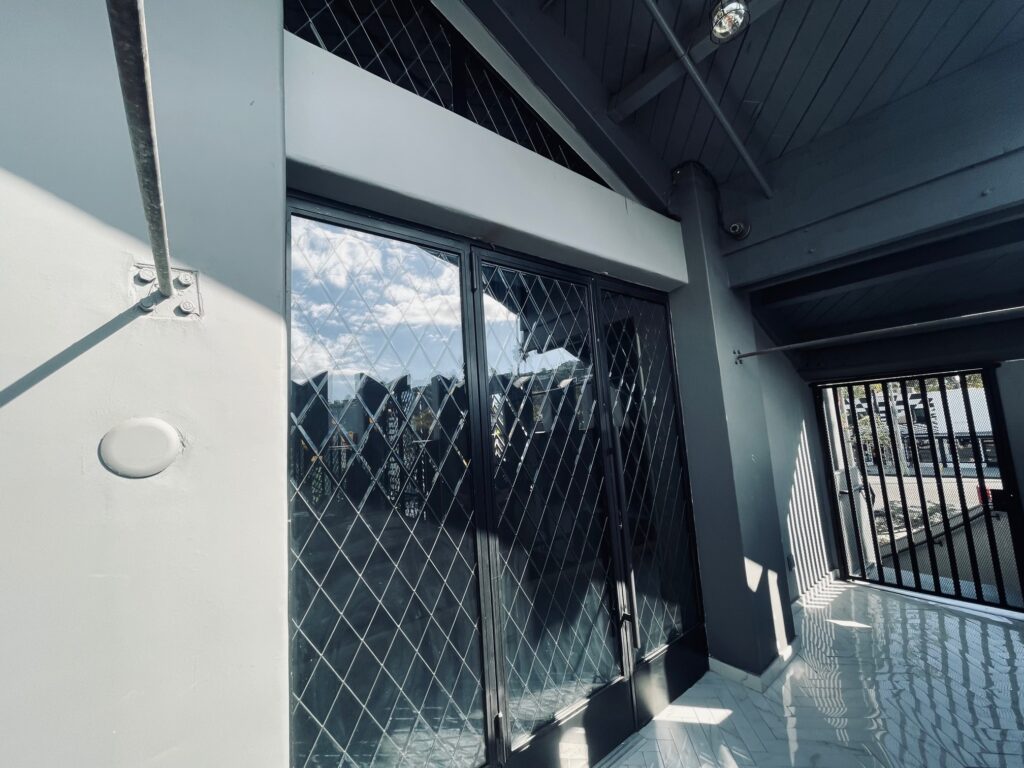 Exterior view of multiple clear leaded glass panels with a classic diamond pattern, set in dark frames, reflecting a cloudy blue sky. The panels form part of a modern building's entrance or facade, featuring light gray walls, a dark gray slatted ceiling, and a reflective light-colored tiled floor, with strong sunlight casting geometric shadows.
