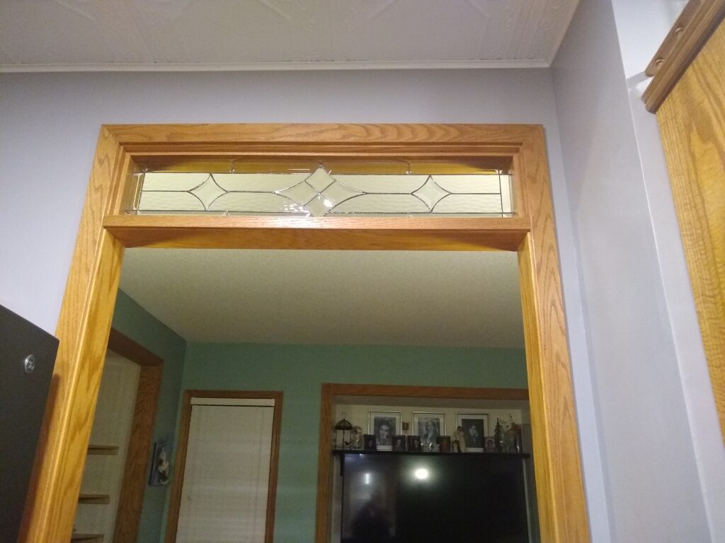A rectangular clear leaded stained glass transom window panel installed above an interior doorway with light oak wood trim. The panel features a central geometric design of clear beveled diamond shapes, flanked by textured clear glass panels and elongated diamond sections, enhancing privacy and light.