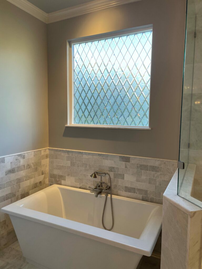 A rectangular clear textured leaded glass window with a repeating diamond pattern, set above a white freestanding tub in a modern bathroom with light gray walls and tiled wainscoting.