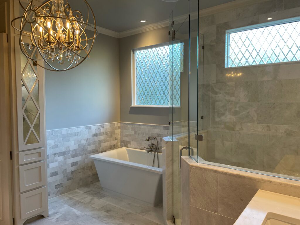 A luxurious modern bathroom featuring a freestanding white bathtub, a glass-enclosed walk-in shower, and marble-look tiles. Two windows, a large horizontal one and a smaller vertical one, are fitted with clear, textured leaded glass panels forming a diamond lattice pattern, providing privacy while allowing abundant natural light. A decorative gold chandelier hangs in the foreground.