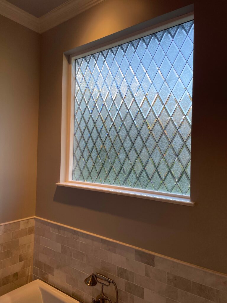 A rectangular leaded glass window in a bathroom, featuring a repeating diamond pattern composed of clear beveled glass and textured glue chip glass, providing privacy. The window is set in a white frame above a light-colored tiled wall and a bathtub.