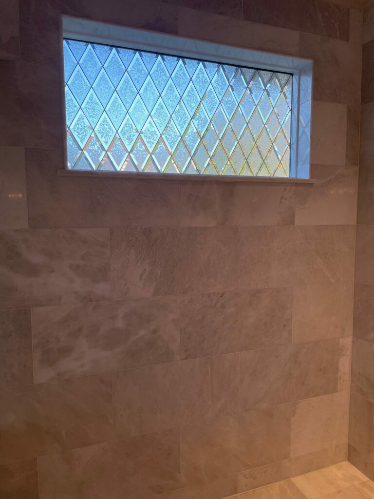A rectangular leaded glass window featuring a uniform diamond pattern. Each diamond pane consists of highly textured, clear glass that subtly refracts light, providing privacy. The window is framed by light trim and set into a wall covered with large rectangular light beige marble-look tiles.