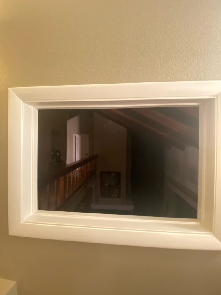 A horizontally oriented, rectangular mirror with a thick, clean white wooden frame, reflecting a dimly lit indoor hallway with a visible wooden staircase, banister, and a distant white door.