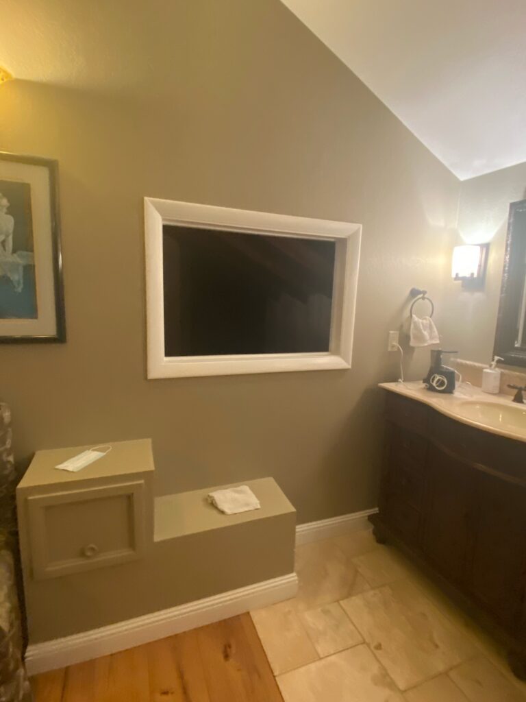 A rectangular clear beveled leaded stained glass privacy panel, featuring a geometric pattern of textured clear glass and faceted bevels, installed in a prominent white frame on a warm beige wall, adjacent to a dark wood bathroom vanity and a wall-mounted sconce.