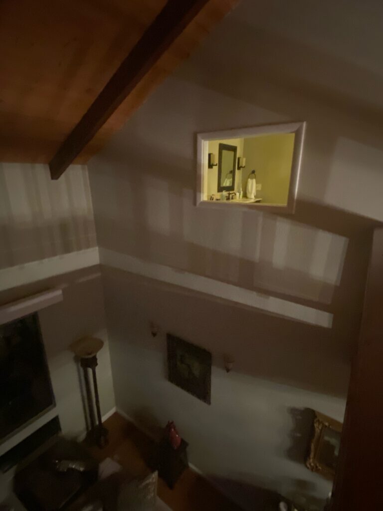 High-angle view inside a split-level home, showing an upper landing with dark wooden ceiling beams and light walls. A rectangular pass-through window opens to a bathroom vanity with a mirror and sink. Below, part of a living area with furniture and wall decor is visible, featuring various light and shadow patterns.