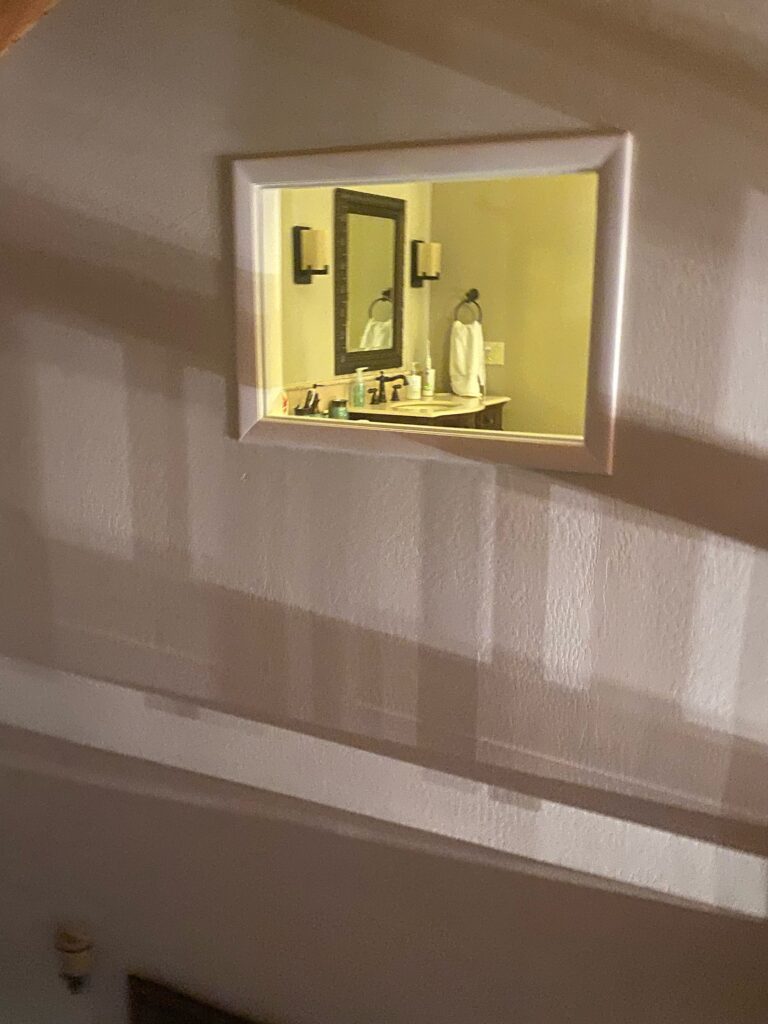 A rectangular, wall-mounted stained glass privacy panel featuring a highly reflective surface, reflecting a bathroom vanity with a sink, a smaller mirror, wall sconces, and a white towel.