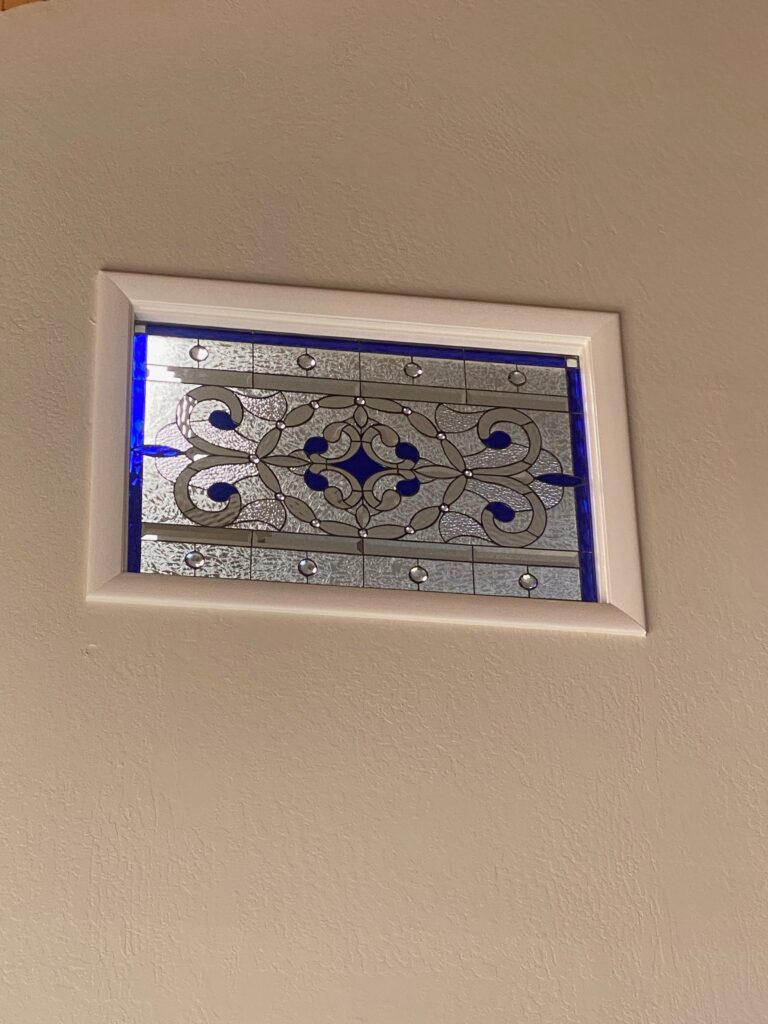 A rectangular leaded stained glass panel with a symmetrical, ornate scroll and floral motif, crafted from clear textured and beveled glass, vibrant sapphire blue accents, and embedded clear bevel jewels, framed in white and set into a textured beige wall.