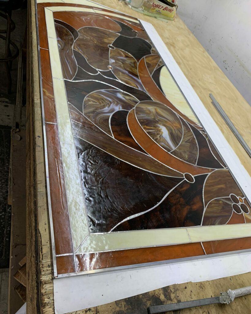 A rectangular stained glass panel featuring an intricate, organic Art Nouveau design. The glass is primarily in warm tones of swirling dark brown, amber, and cream, with elegant curved lines and circular accents. A border of lighter cream and solid rust-brown glass frames the central abstract pattern, all joined by silver lead lines.