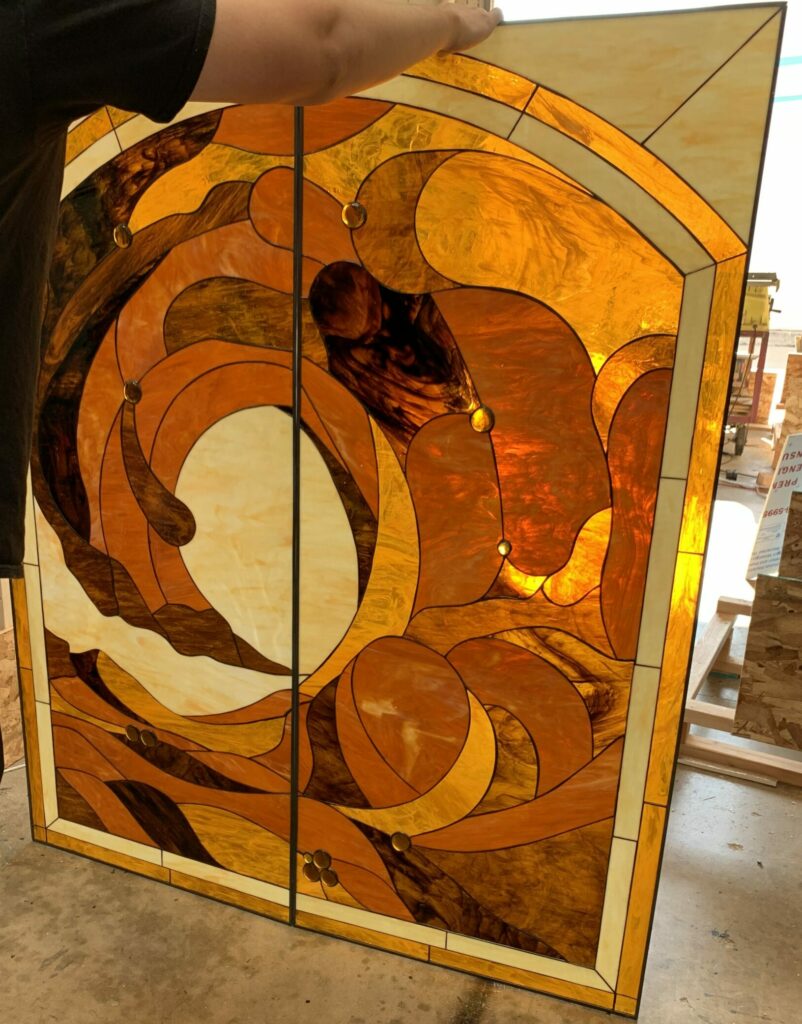 A person holds a large, rectangular stained glass artwork composed of two vertical panels. The artwork features an abstract, organic design in varying shades of amber, gold, and dark brown, with lighter cream-colored sections. Several round glass jewels are embedded within the flowing patterns. Dark lead lines delineate the shapes, and a light-colored border frames the entire piece.
