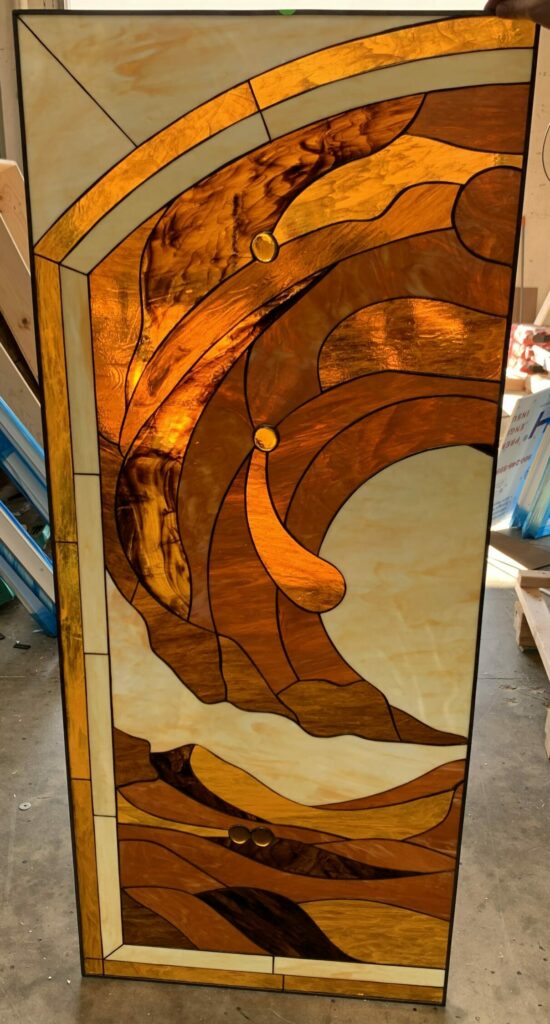 A rectangular leaded stained glass panel with an abstract, organic design resembling flowing waves. It features various shades of amber, golden brown, and light cream-colored art glass, accented with two round amber glass jewels.