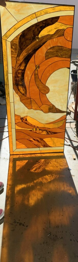 A tall, rectangular stained glass panel featuring an abstract design of flowing, organic shapes in various shades of molten amber, golden orange, and dark brown, accented with small gold glass jewels. A person holds the panel upright in a sunlit workshop, casting vivid orange and yellow patterns onto the concrete floor.