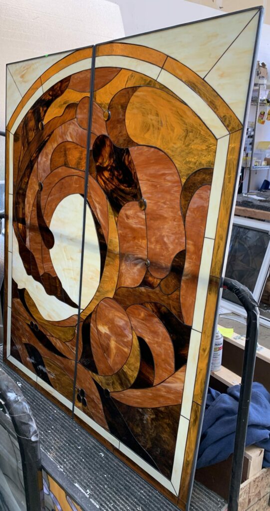 A large, two-panel arched stained glass window featuring an abstract, flowing design in amber, brown, and cream-colored opaque glass, with integrated circular glass accents.