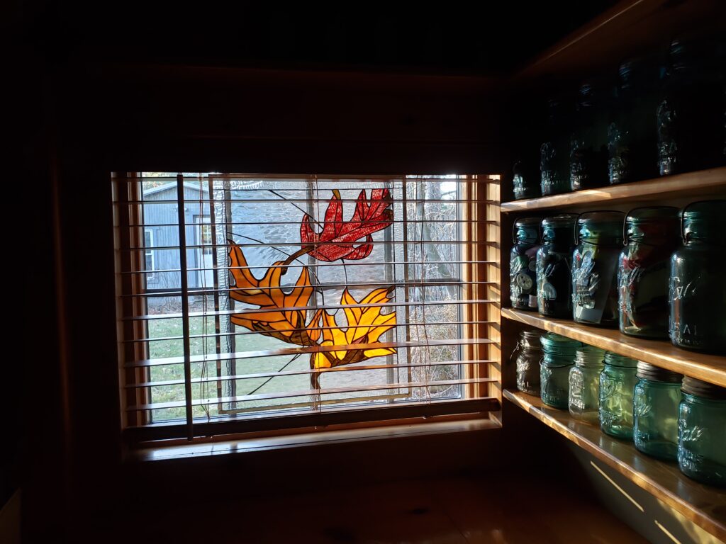 A rectangular leaded stained glass window panel featuring three autumn leaves—one vibrant red and two warm amber/orange—installed within a window. The window is fitted with horizontal wooden blinds, and sunlight streams through the stained glass. To the right, wooden shelves display several vintage blue glass canning jars. Outside the window, a shed and trees are visible.