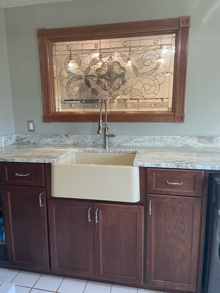 A rectangular leaded stained glass window panel installed above a farmhouse sink in a laundry room or kitchen. The clear glass panel features an intricate Victorian-inspired scroll and floral design, combining various textured and beveled glass pieces. It is set within a robust, ornate dark wood frame with fluted columns and carved corner rosettes.