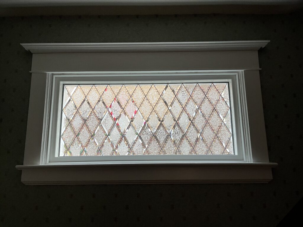 A rectangular leaded glass window set into white interior trim, featuring an all-clear diamond lattice design. The individual diamond panes alternate between clear beveled glass and clear heavily textured or crackle glass, providing privacy while allowing ample natural light. A red brick wall is partially visible outside through the glass, and the surrounding room has dark patterned wallpaper.