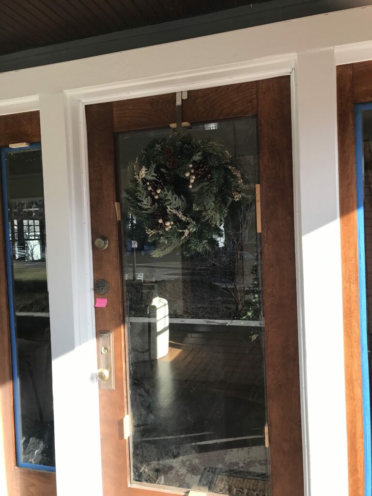 A brown wooden front entry door with a large clear glass panel and two clear glass sidelights, all framed by clean white trim. A holiday wreath with pinecones and shimmering accents hangs on the main door, and blue painter's tape is visible on the left sidelight, indicating ongoing work.