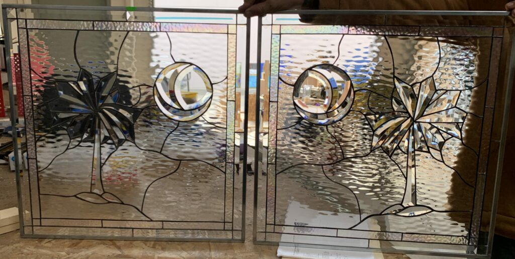 Two identical clear leaded stained glass panels, each featuring a central beveled glass sunburst motif and an offset beveled glass crescent moon within a clear circular outline. The background is composed of clear, heavily textured water glass, all framed by clear beveled border pieces. A person's hands hold the panels from the top in a workshop setting.