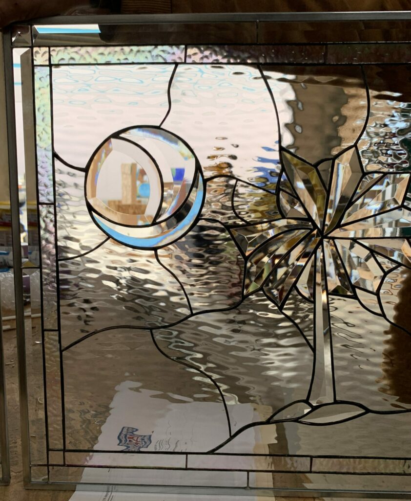 A rectangular clear leaded stained glass panel with a contemporary design. It features a large, multi-faceted beveled glass starburst on the right, and a circular beveled element with a subtle iridescent crescent on the left, all set against a textured ripple glass background. The panel is held by a person's hand in a workshop.