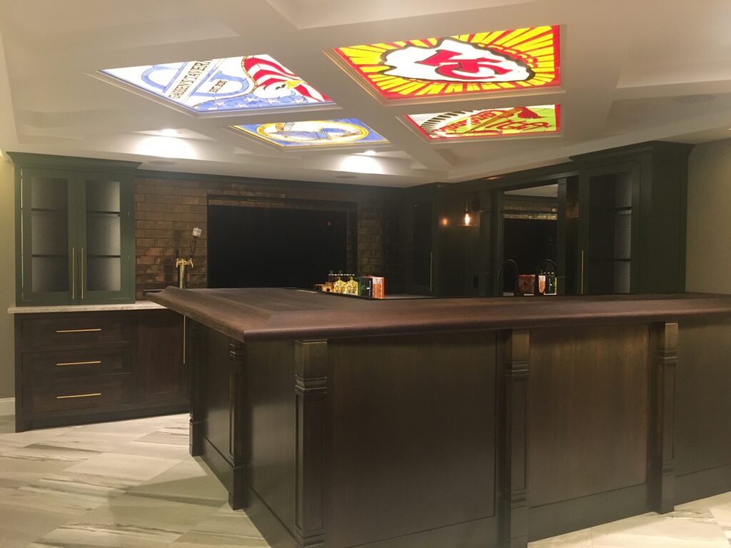 A luxurious custom home bar featuring dark wood cabinetry, a rich wooden countertop, and four illuminated stained glass panels recessed into the ceiling. The panels display various sports team logos, including the Kansas City Chiefs, and a personalized 'Gassen's Tavern' sign.