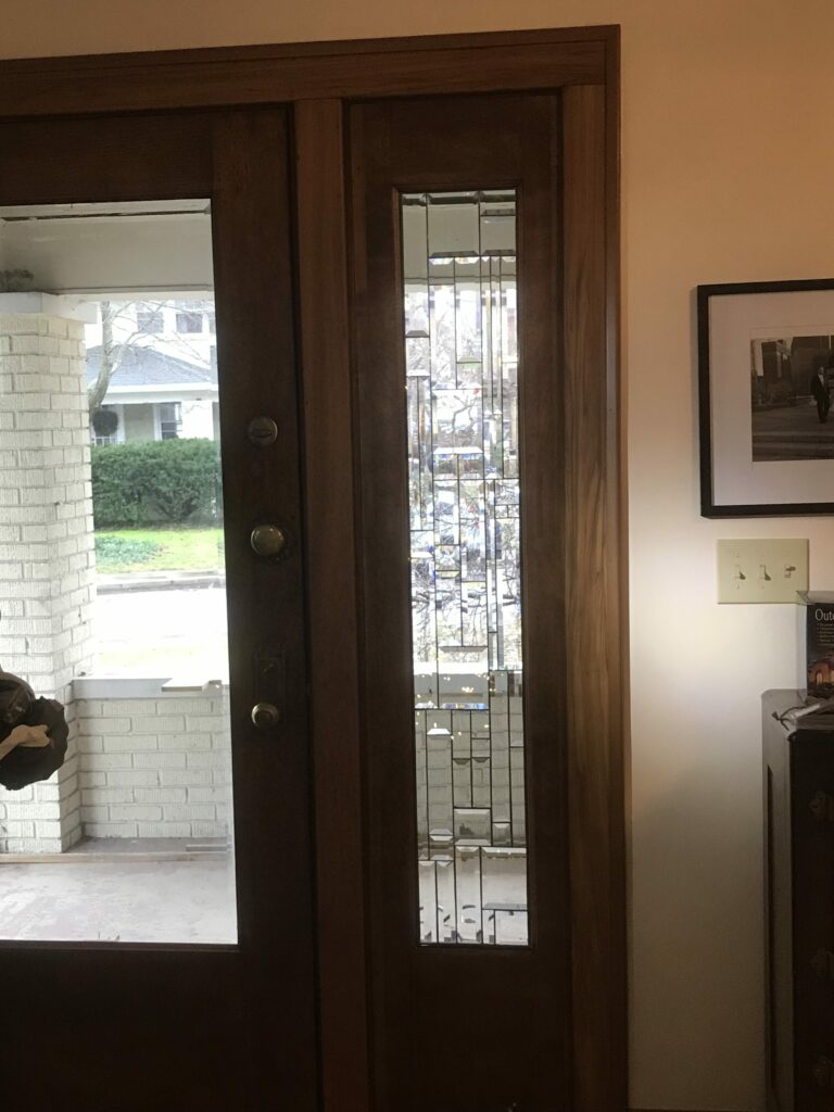A rectangular, clear beveled and textured leaded stained glass panel showcasing an abstract geometric design, installed in a dark wooden entryway sidelight.