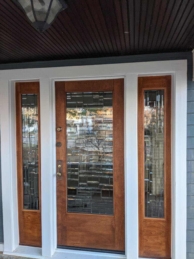 Front exterior view of a wooden entry door flanked by two sidelights, each featuring rectangular leaded glass panels with a modern geometric pattern of clear beveled and darker gray glass blocks. The main door has brass hardware, and the entire unit is set in white trim against blue house siding.