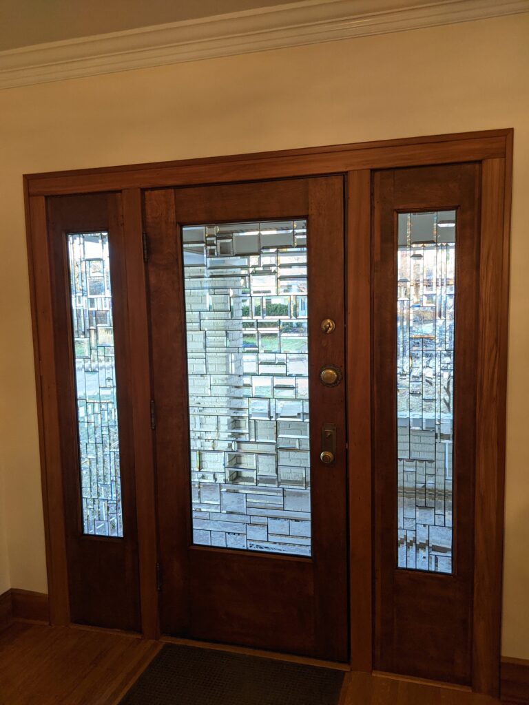 A wooden entryway featuring a main door and two narrow sidelights, all fitted with clear, textured, and beveled leaded glass panels in a geometric, rectilinear pattern. The glass provides privacy while allowing natural light, and the main door has brass-toned hardware.
