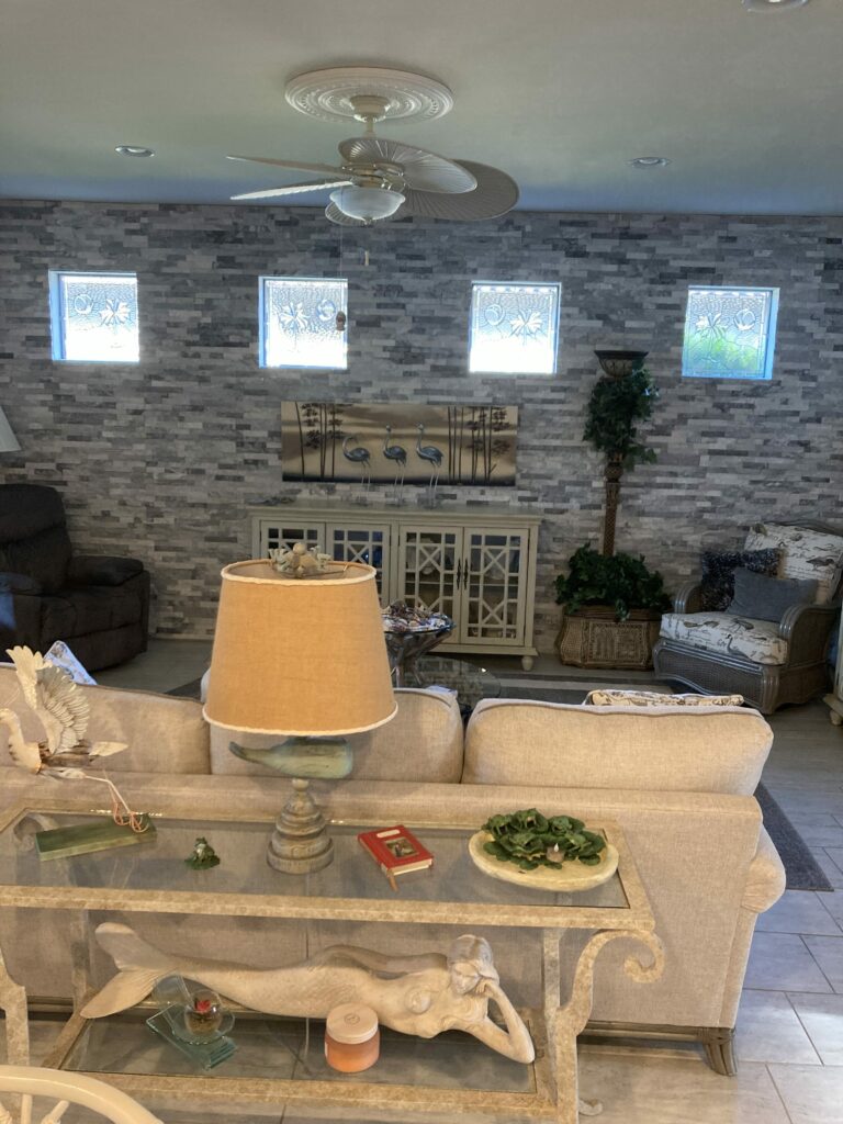 Four clear beveled glass transom windows installed high on a gray stone-patterned wall in a living room. Each rectangular window features a central starburst design with radiating beveled cuts, surrounded by clear textured glass and accented by a prominent circular bevel. The glass allows light through while maintaining privacy.
