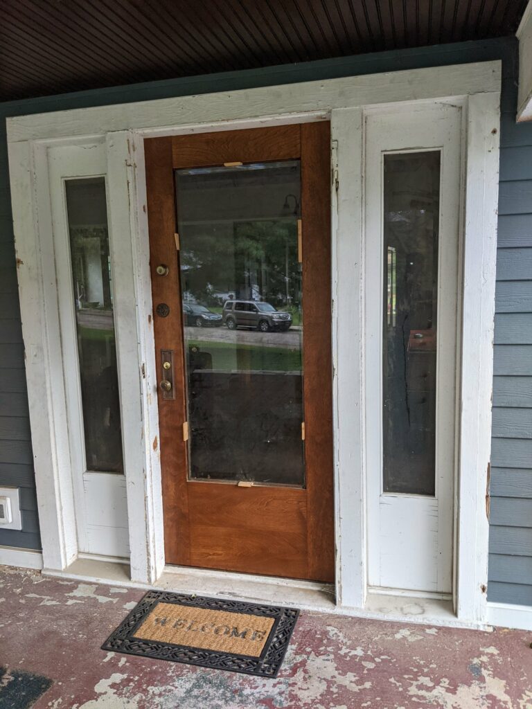 Exterior view of a distressed white-framed front door entrance, featuring a wooden door with clear glass panels and two clear glass sidelights. A 'WELCOME' mat rests on the worn porch floor.