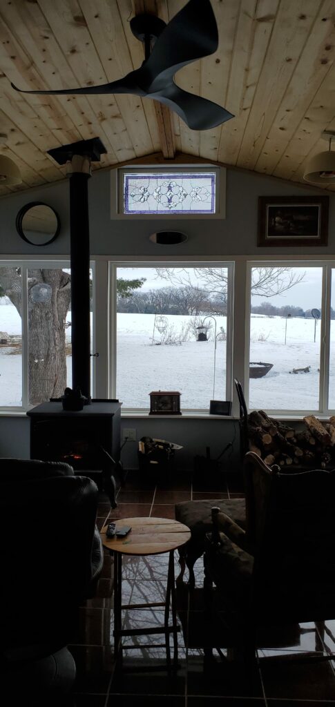 A rectangular stained glass transom panel with a clear textured geometric pattern, featuring a lavender border and subtle purple accents. It is positioned above three large windows, looking out onto a snow-covered field, within a room that has a natural wood ceiling and a wood-burning stove below.