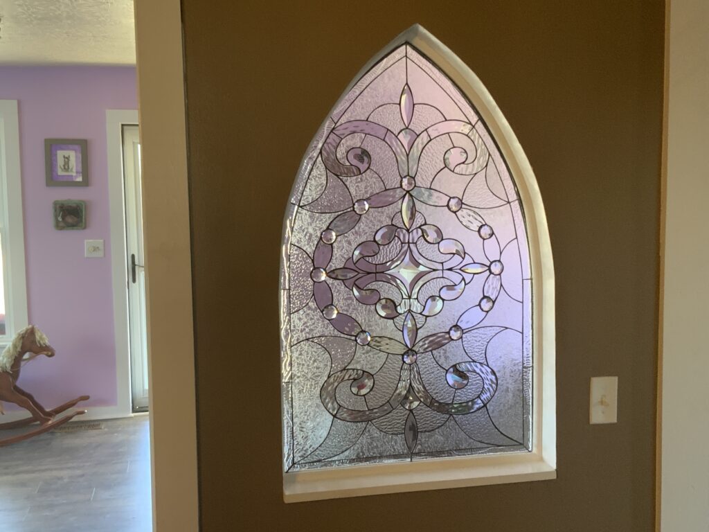 An ornate, leaded stained glass panel in a Gothic arch shape, featuring an intricate abstract design of swirling scrollwork, floral-inspired patterns, textured clear glass, and numerous sparkling clear beveled jewels. The panel is installed in an interior wall between a brown-painted room and a light purple-painted room.