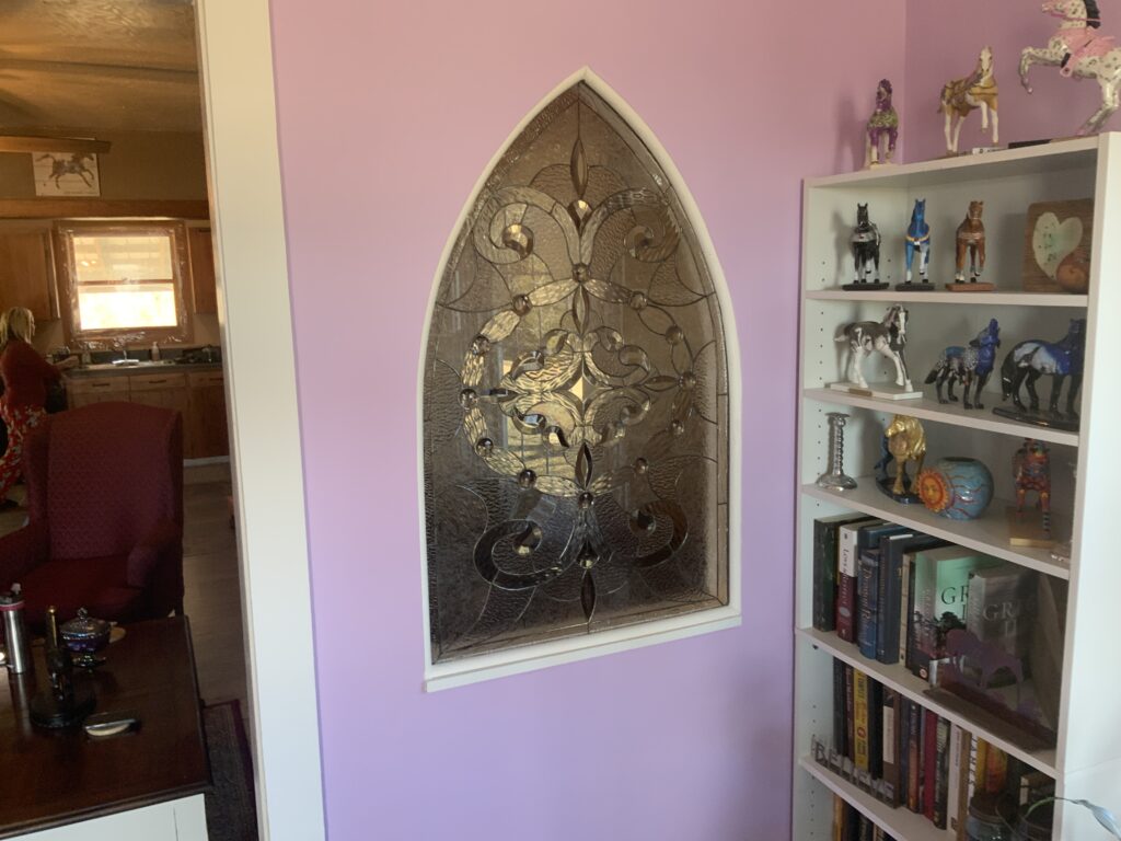 An arched clear leaded stained glass panel mounted on a light purple wall, featuring an intricate design of scrolling patterns and stylized fleur-de-lis motifs with circular bevels, crafted from various clear textured and beveled glass types.