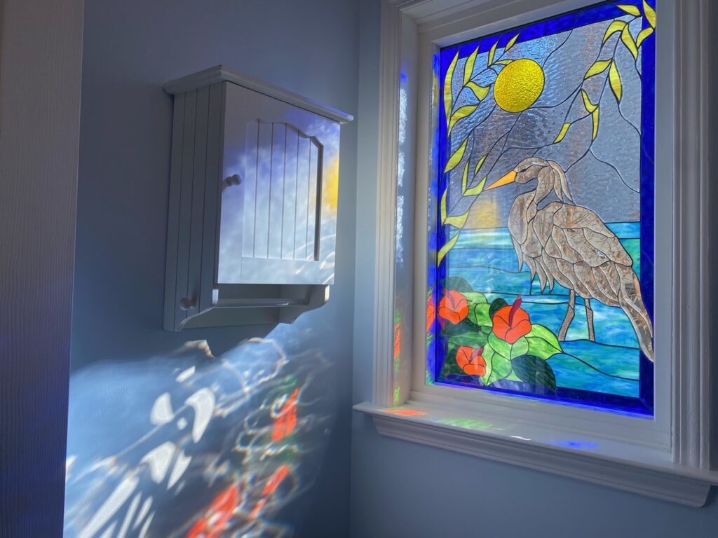 A rectangular leaded stained glass window depicting a large, detailed brown and tan heron standing in blue water with green leaves and vibrant orange hibiscus flowers. A golden yellow sun shines against a light blue textured sky with yellow leafy branches. The window, set in a white frame, is illuminated by bright sunlight, casting colorful, dappled reflections onto an adjacent light blue wall and a white wall-mounted cabinet.