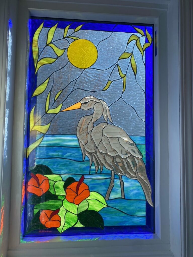 A rectangular stained glass window panel depicting a majestic grey heron with an orange beak standing in blue waters, surrounded by vibrant orange hibiscus flowers and rich green leaves. A bright yellow sun and delicate green foliage are set against a textured light blue sky, all bordered by a striking deep blue glass frame.