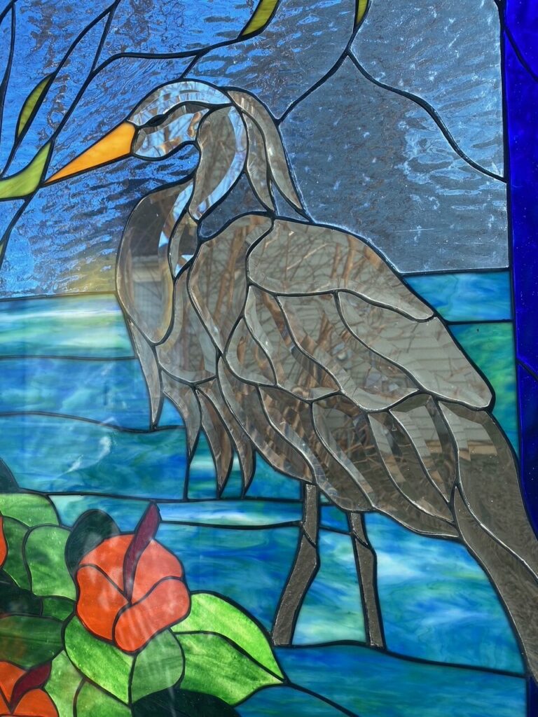 A rectangular leaded stained glass panel featuring a tall, slender grey heron with an orange beak, standing in variegated blue and turquoise water. The heron's body is crafted from various clear and textured grey glass pieces, subtly reflecting tree branches. In the lower left, vibrant red-orange hibiscus flowers with green leaves are prominently displayed. The background transitions from a deep blue sky to a mottled lighter blue, with abstract yellow-green foliage on the left and a solid dark blue border on the right.