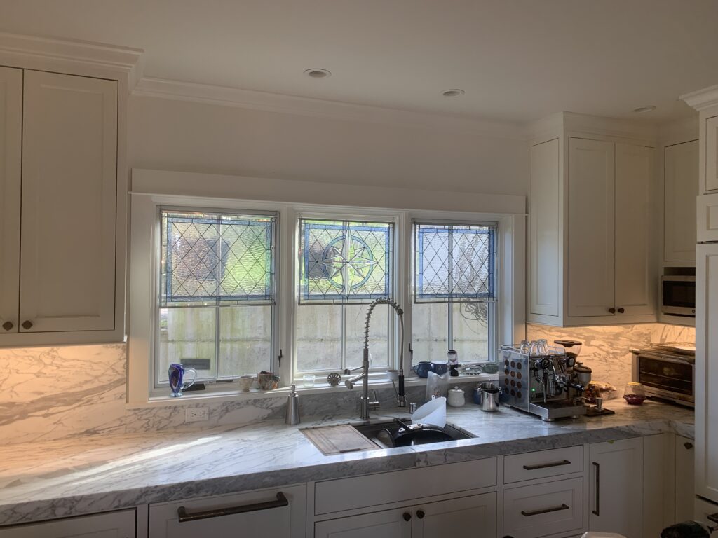 Three rectangular kitchen windows are fitted with custom leaded stained glass panels. The two outer panels feature a clear textured diamond pattern with blue glass borders, while the central panel prominently displays a compass rose medallion in its center, surrounded by the same diamond and blue border design. The windows are situated above a marble countertop with a sink and a modern espresso machine, flanked by white shaker-style kitchen cabinetry.