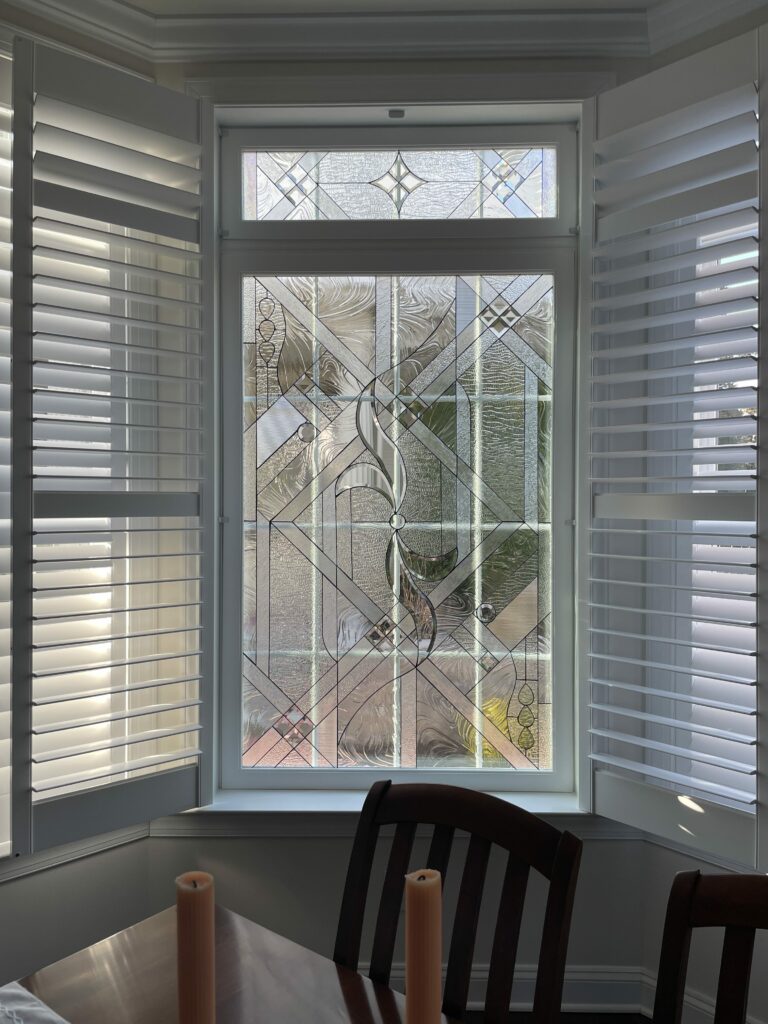 A rectangular leaded stained glass window, consisting of a main panel and a smaller transom above, set within a white frame. The clear glass design features an intricate abstract geometric pattern with numerous beveled accents, various textured glass (e.g., ripple, hammered, seedy), and stylized organic motifs including a central scroll and teardrop shapes on the sides. White plantation shutters are partially open on either side, revealing natural light. A dark wooden dining table and chairs are partially visible in the foreground.