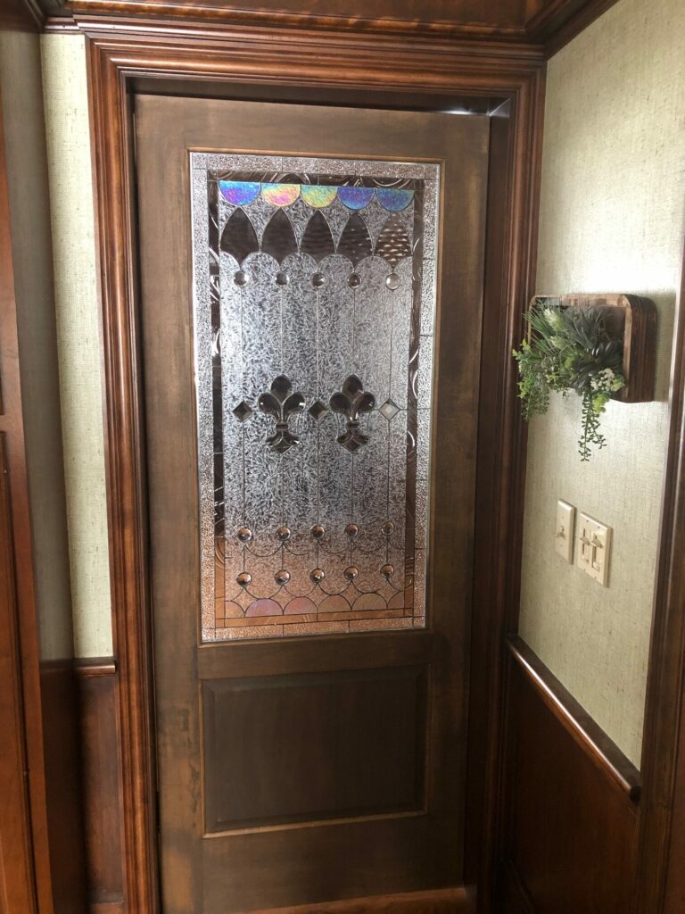 A full-length dark brown wooden door featuring a rectangular leaded stained glass insert. The central panel is made of heavily textured clear glass, accented by two prominent dark amber Fleur-de-lis motifs with clear beveled outlining. The top border of the glass panel displays iridescent blue, purple, and green semicircle pieces above dark, scalloped glass. The bottom border includes clear glass cabochons and additional light amber textured glass. The door is set within rich wooden trim next to a light-colored, textured wall, which features a wooden wall planter with artificial greenery and light switches.