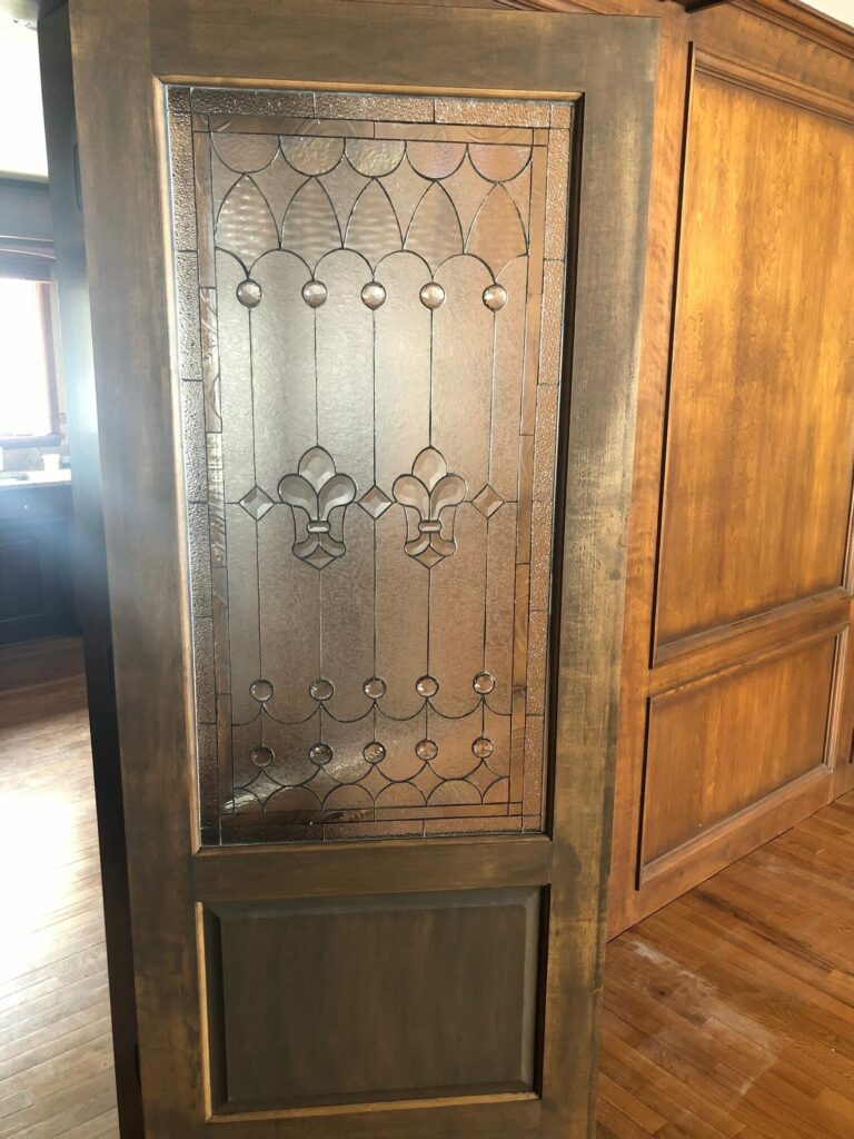 A rectangular clear leaded stained glass panel featuring prominent Fleur-de-lis motifs, intricate geometric patterns, textured glass, and round clear bevels, set within a dark stained wooden interior door.