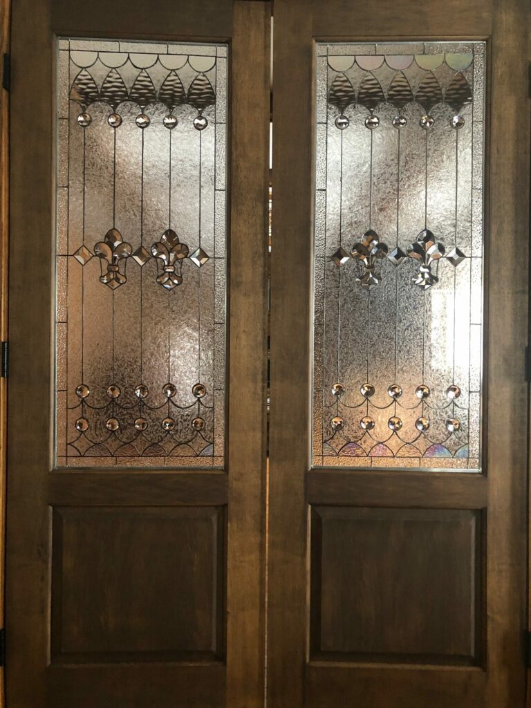 A pair of rich brown wooden double doors, each featuring a rectangular leaded stained glass panel. The panels are crafted with clear textured glass for privacy, accented by brilliant beveled glass Fleur-de-lis designs, round beveled jewels, and geometric patterns.